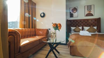 Une chambre d’hôtel avec un canapé marron, une table basse en verre, un lit avec une tête de lit marron et un mur orné de deux œuvres d’art encadrées.