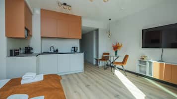 A modern hotel room with a bed, a kitchenette, a dining area with a table and chairs, and a TV mounted on the wall.