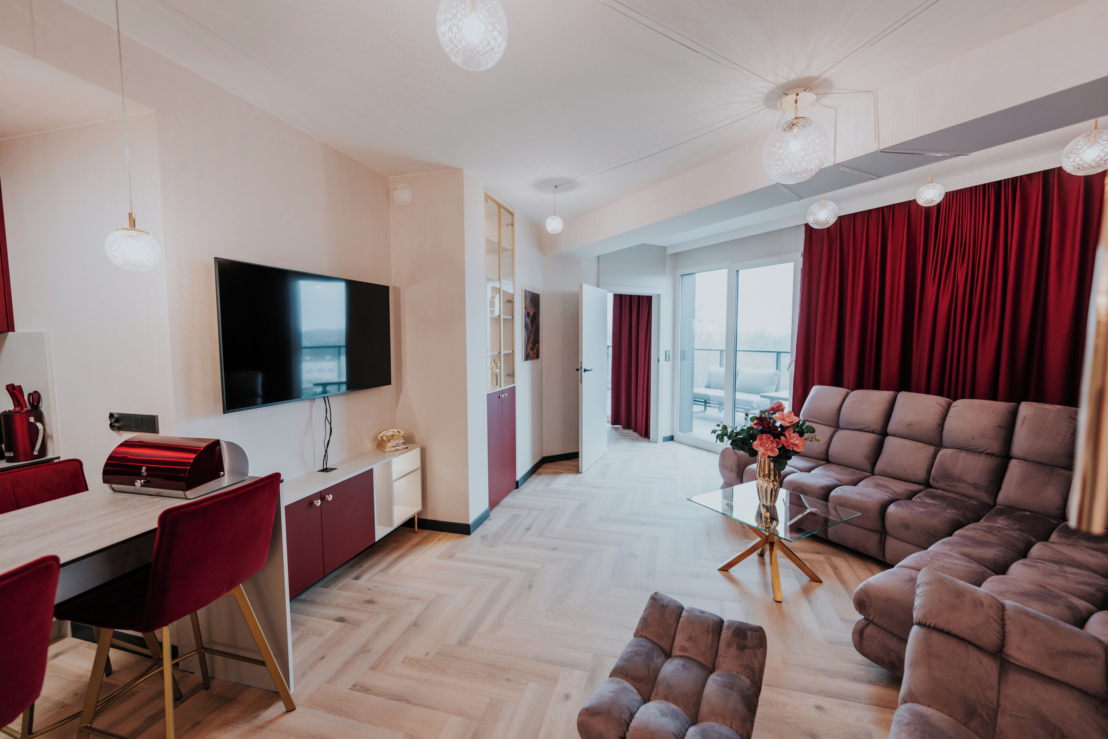 A modern living room with a flat-screen TV, a sofa, a dining area with red chairs, and a large window with curtains.