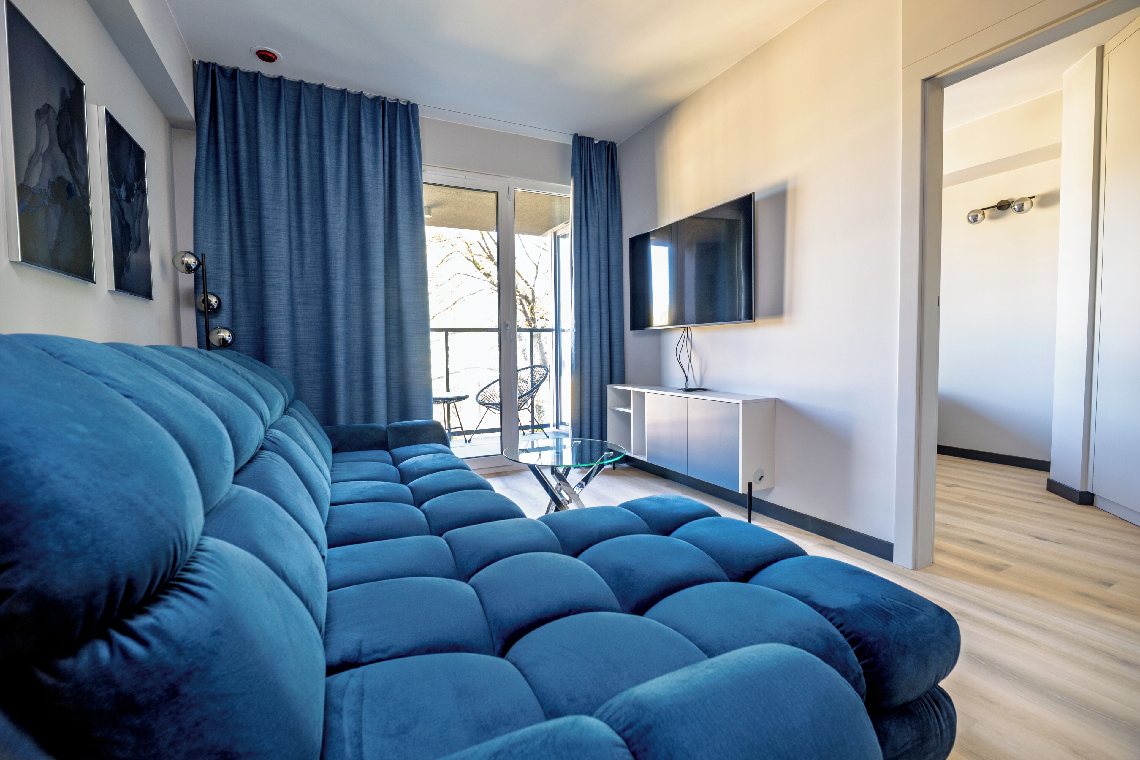 A modern living room with a large blue sofa, a flat-screen TV mounted on the wall, and a glass dining table.