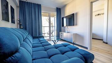A modern living room with a large blue sofa, a flat-screen TV mounted on the wall, and a glass dining table.