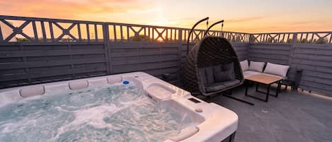 A hot tub on a rooftop patio with a wooden fence, a wicker swing chair, and a table with cushions.
