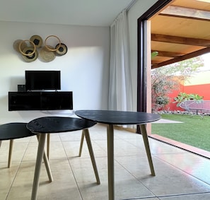 A modern living space with a flat-screen TV, two black tables with wooden legs, and a large window with curtains. - A two-bedroom villa with saltwater swimming pool, sun terrace, BBQ and gardens (Corralejo)