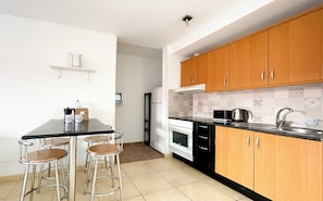 A kitchen with a black countertop, wooden cabinets, a white refrigerator, and a microwave. There are two bar stools and a small table with a water bottle and a box. - A two-bedroom villa with saltwater swimming pool, sun terrace, BBQ and gardens (Corralejo)