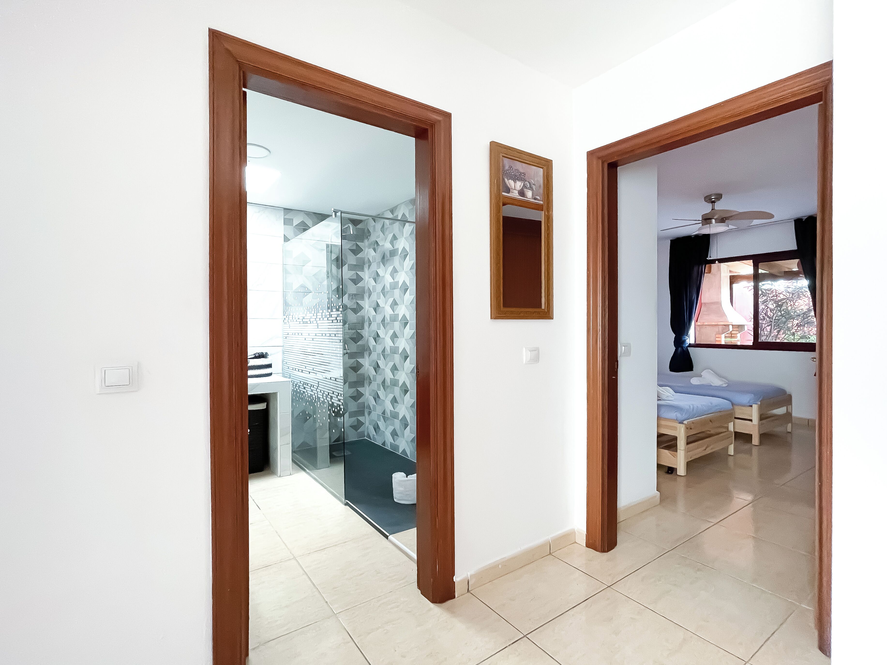 A hallway with a glass door leading to a bathroom, a bed with a ceiling fan, and a window with curtains.