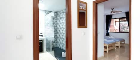 A hallway with a glass door leading to a bathroom, a bed with a ceiling fan, and a window with curtains.