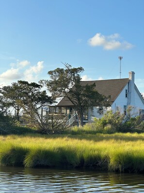 A white house with a chimney, surrounded by grass and trees, near a body of water.