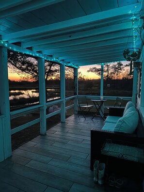 Un espace extérieur abrité, avec des places assises, offrant une vue sur un coucher de soleil se reflétant sur une étendue d’eau, un sol en bois et une table avec des chaises.
