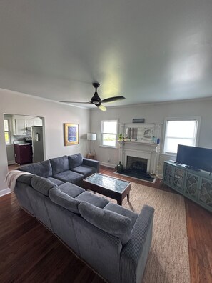Una sala de estar con sofá modular, chimenea, televisor y una cocina al fondo.