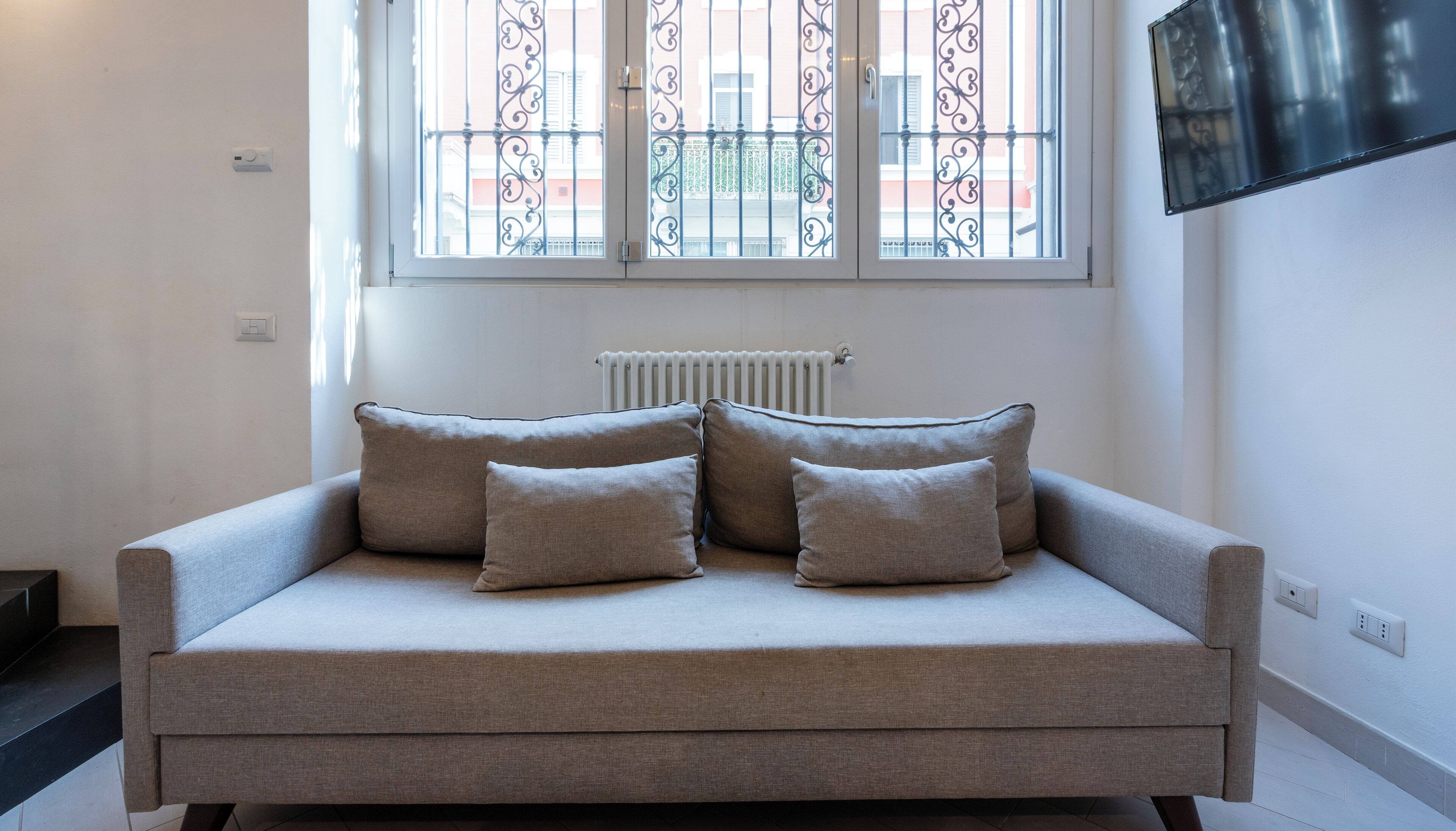 Un sofá de color neutro con cuatro cojines en una habitación que tiene un radiador, una ventana con barandillas decorativas y un televisor montado en la pared.