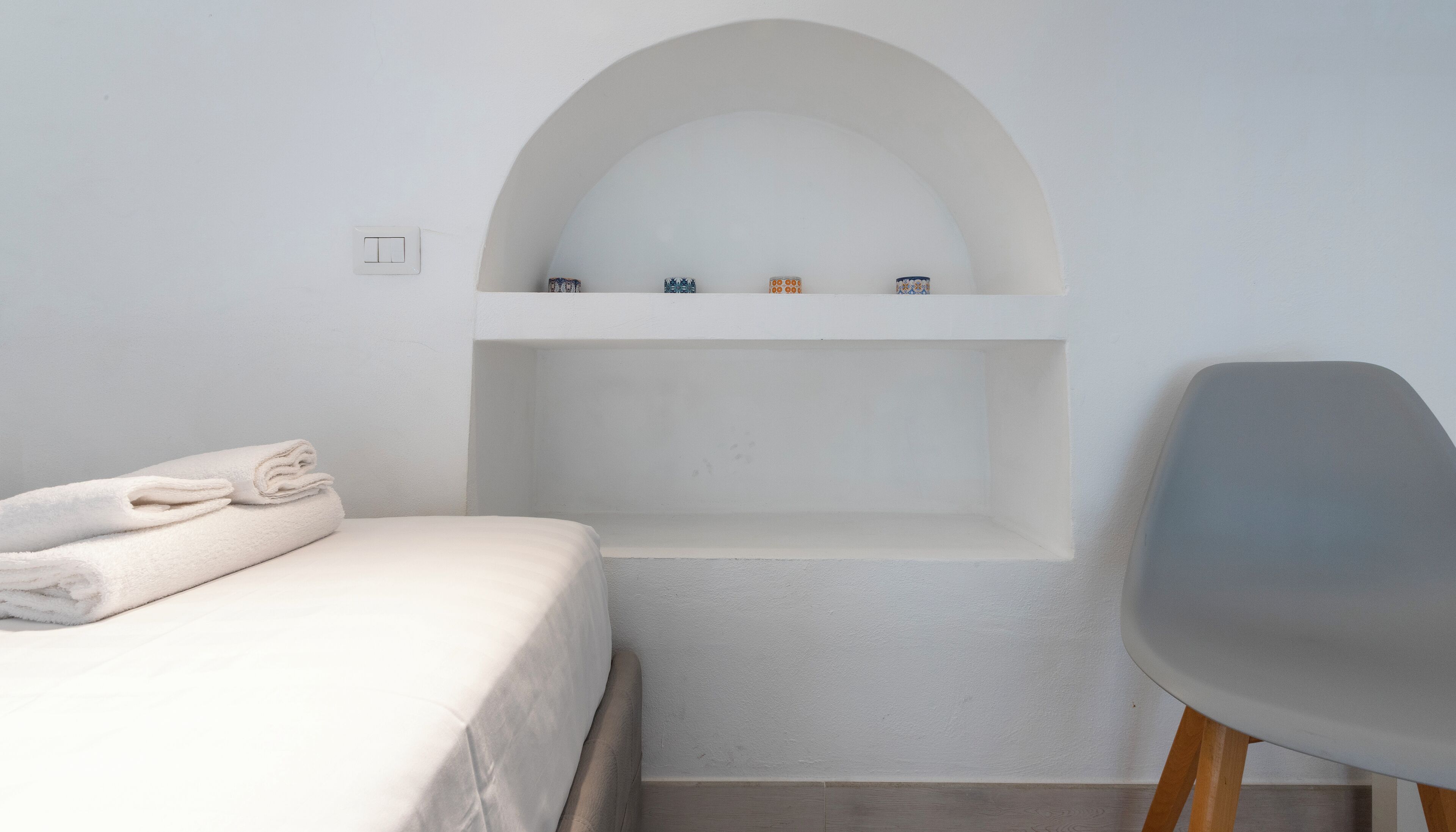 Habitación minimalista con cama, una silla y estante empotrado con objetos decorativos.