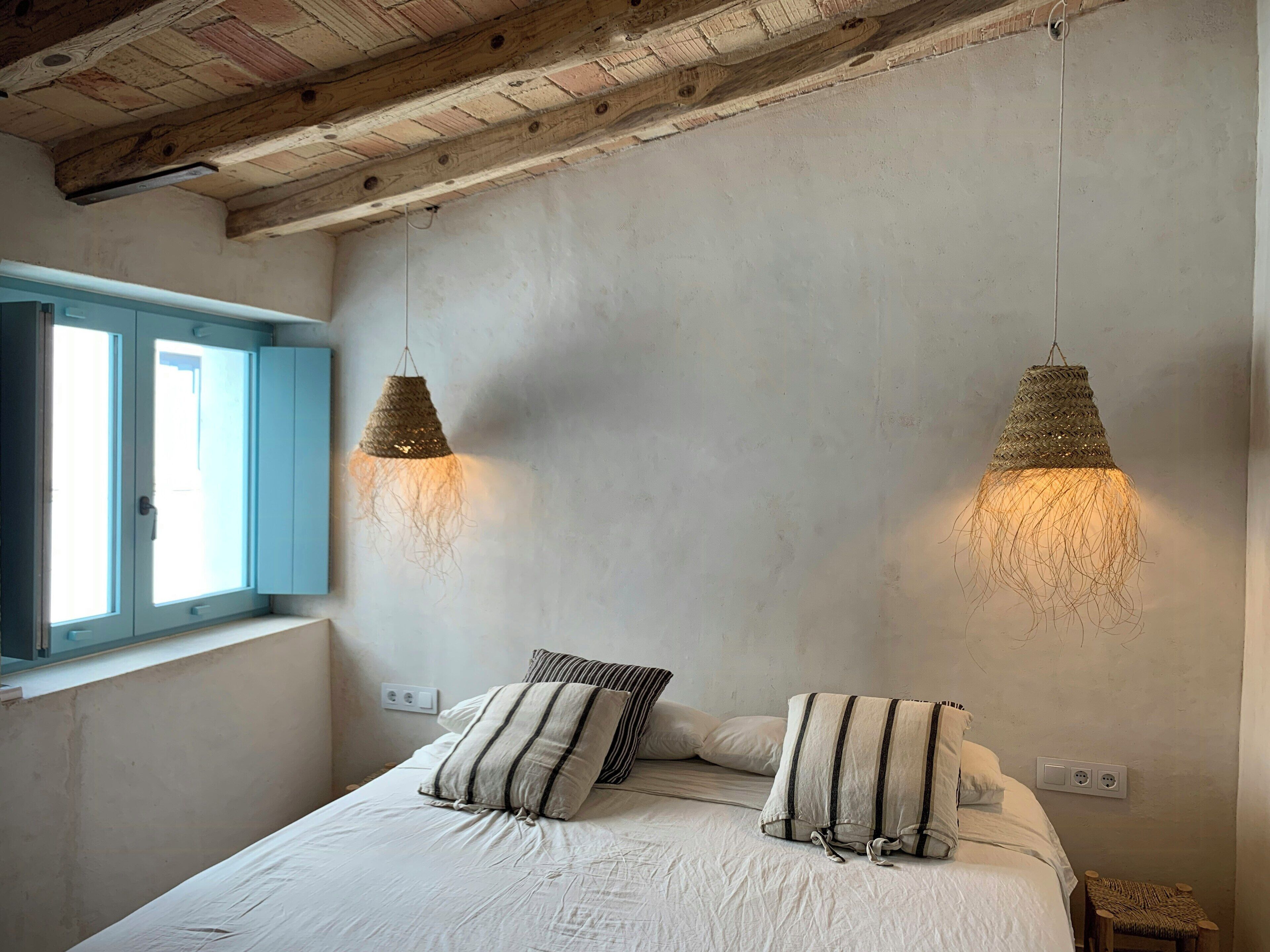 A bedroom with a bed, two hanging lamps, a window with blue shutters, and a wall socket.