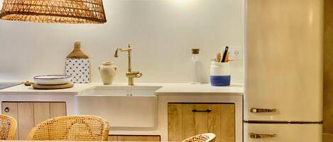 A kitchen with a white countertop, a sink, a refrigerator, wicker chairs, and a hanging light fixture.