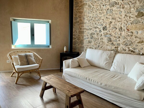 Um quarto com parede de pedra, um sofá branco, uma cadeira de vime, um banco de madeira e uma pequena mesa.
