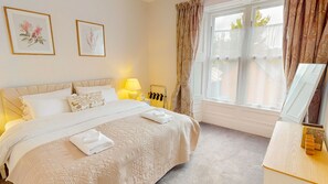 A hotel bedroom with a large bed, a bedside table with a lamp, a mirror, and two framed floral artworks on the wall. - The Twinflower - townhouse in the heart of Grantown on Spey (Grantown-on-Spey)