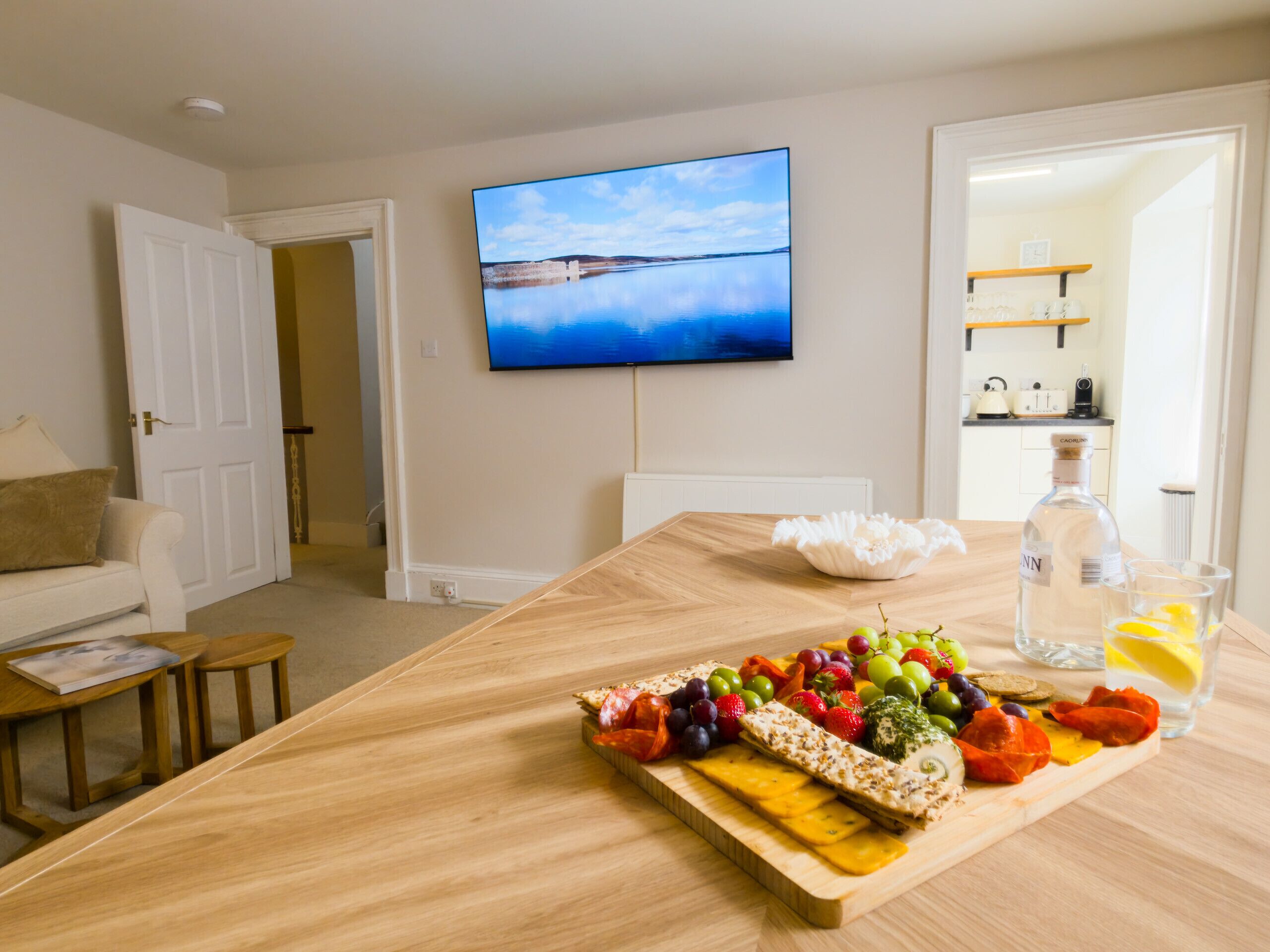 A dining area with a wooden table set with a fruit platter, a bottle of liquor, and glasses. A television is mounted on the wall above the table.