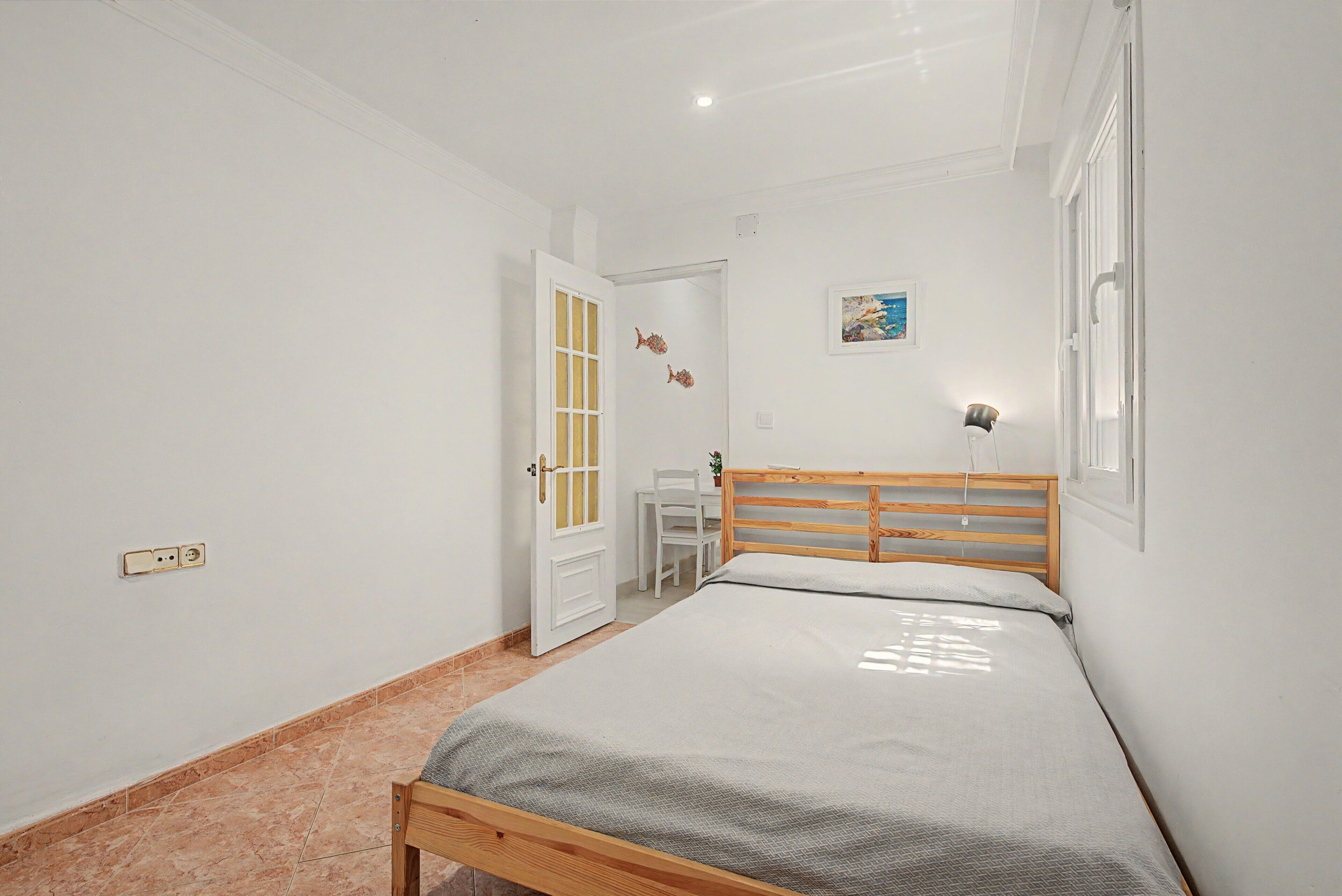 Quarto com cama de madeira, porta branca, mesa pequena e luminária fixada na parede.