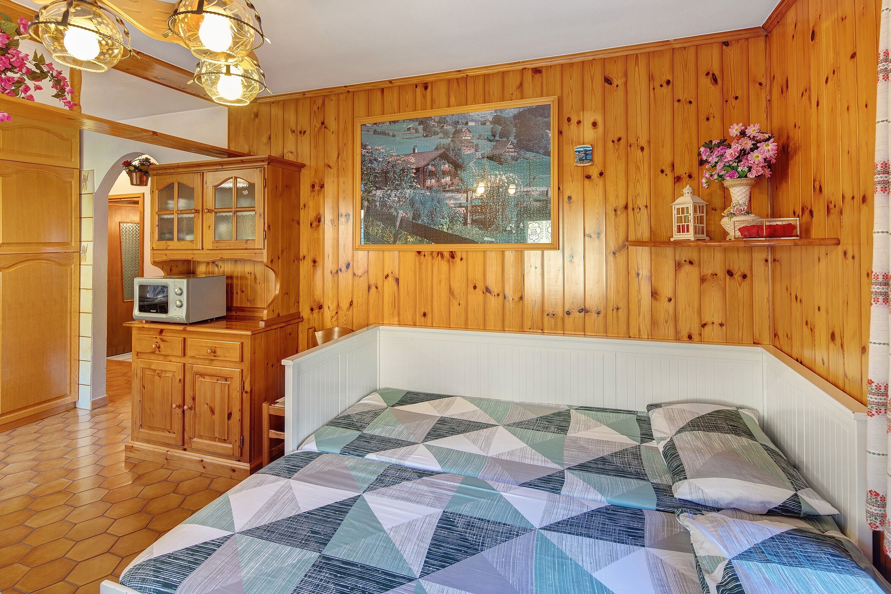 A bedroom with wooden paneling, a bed with a geometric-patterned quilt, a painting of a landscape, and a small shelf with decorative items.