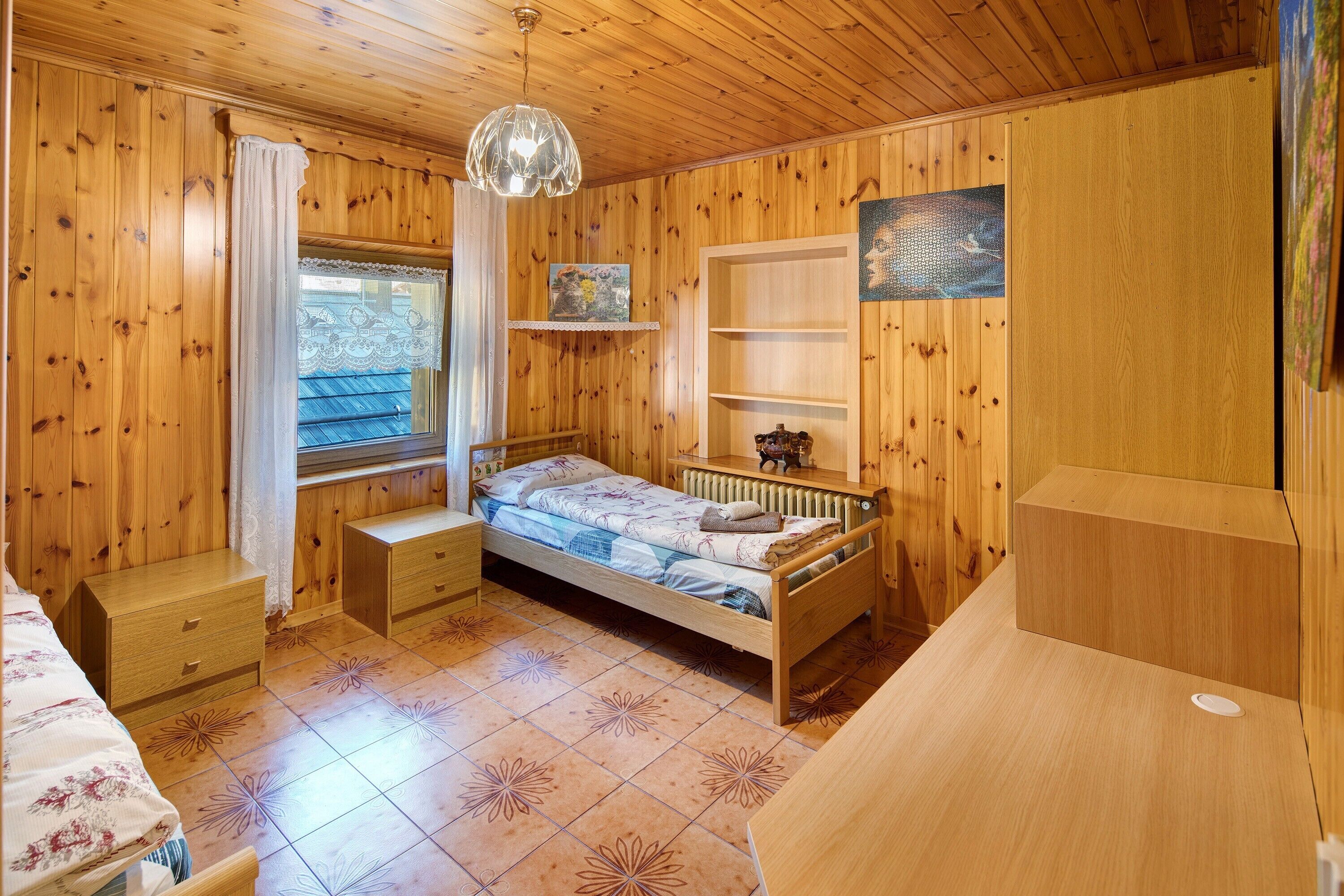 Una stanza in legno con un letto, una finestra con tende, un termosifone e uno scaffale con oggetti decorativi.