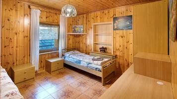 Una stanza in legno con un letto, una finestra con tende, un termosifone e uno scaffale con oggetti decorativi.