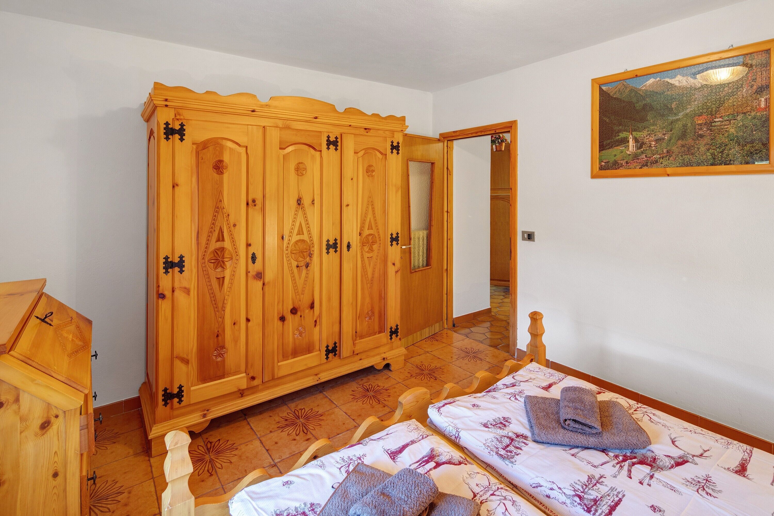 A room with a wooden wardrobe, a bed with patterned bedding, and a painting on the wall.