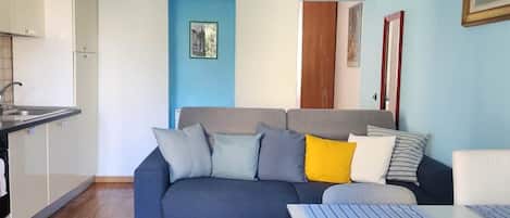 A compact living space with a kitchenette, a sofa with cushions, a dining table, and a bed.