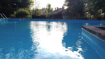 A clear swimming pool with a ladder on the left, surrounded by greenery and buildings in the background.