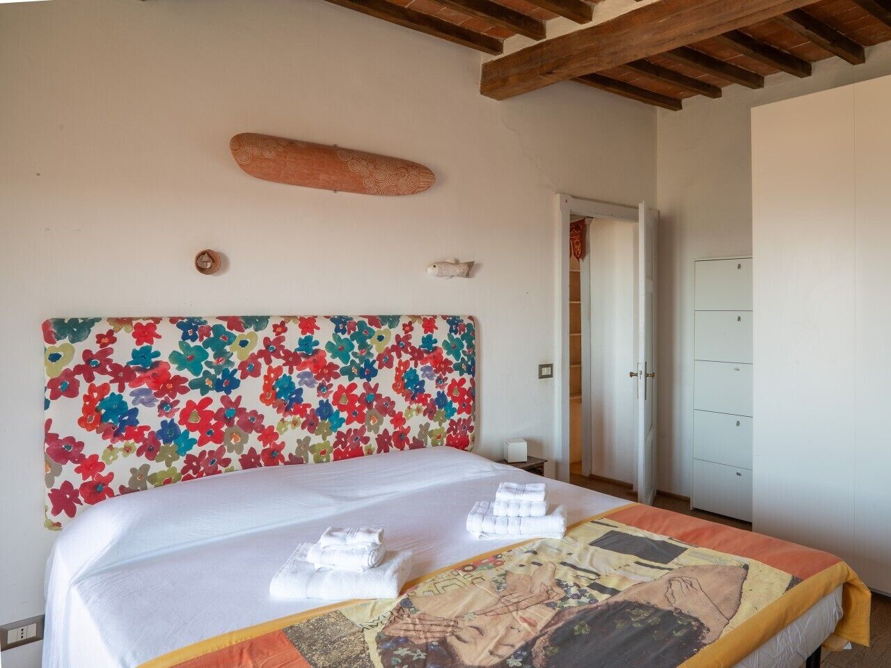 Ein Hotelzimmer mit einem großen Bett, einem farbenfrohen Kopfteil, einer Holzbalkendecke und einem weißen Kleiderschrank.