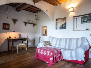 A cozy room with a wooden floor, a striped sofa, a wicker chair, a desk with a lamp, and wall-mounted artwork.