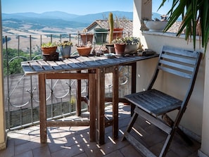 A wooden table and chair on a balcony with potted plants and a view of the countryside.