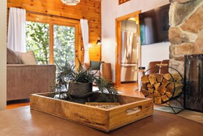 A wooden table with a decorative plant arrangement, a stone fireplace, and a modern kitchen in the background. - Luxury 4BR Mountain Cabin for 8 - Stargazing Dome, Hot Tub & Game Lounge (Harpers Ferry)