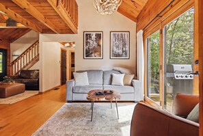 Ein gemütliches Wohnzimmer mit Holzboden, einem grauen Sofa, einem Couchtisch und einem großen Fenster mit einem Grill davor.