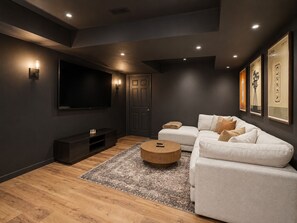 Ein modernes Wohnzimmer mit einem großen Flachbildfernseher, einem dunklen Holzboden, einem gemusterten Teppich und einem weißen Sofa mit vielen Kissen.