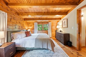 A wooden-paneled bedroom with a bed, bedside table, dresser, and a window with curtains. - Luxury 4BR Mountain Cabin for 8 - Stargazing Dome, Hot Tub & Game Lounge (Harpers Ferry)