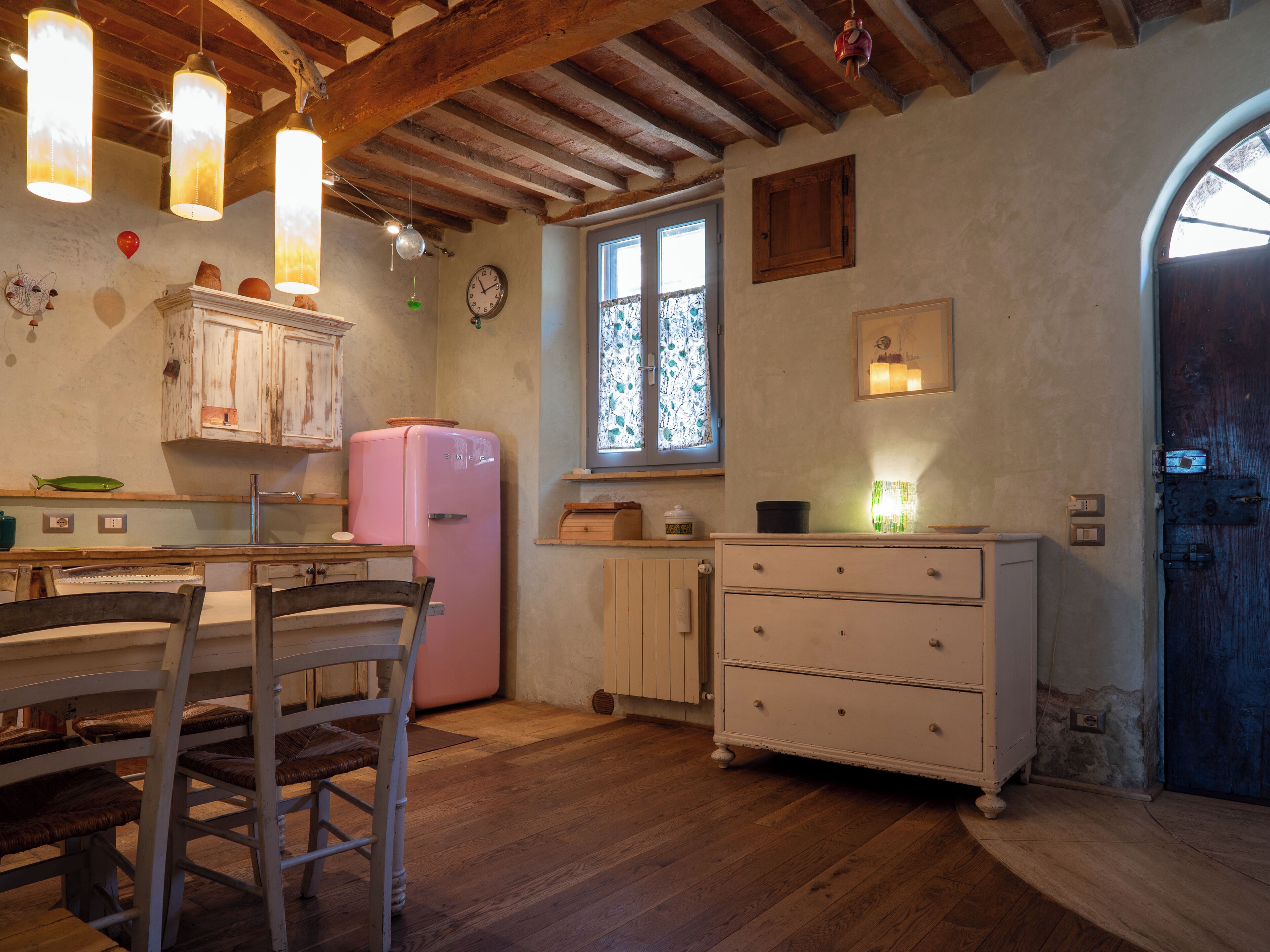 A rustic kitchen with a wooden table, chairs, a pink refrigerator, and a window with patterned curtains.