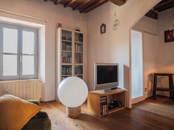 A room with a television on a wooden stand, a bookshelf, and a window with blinds. - Nido con vista (Radicondoli)
