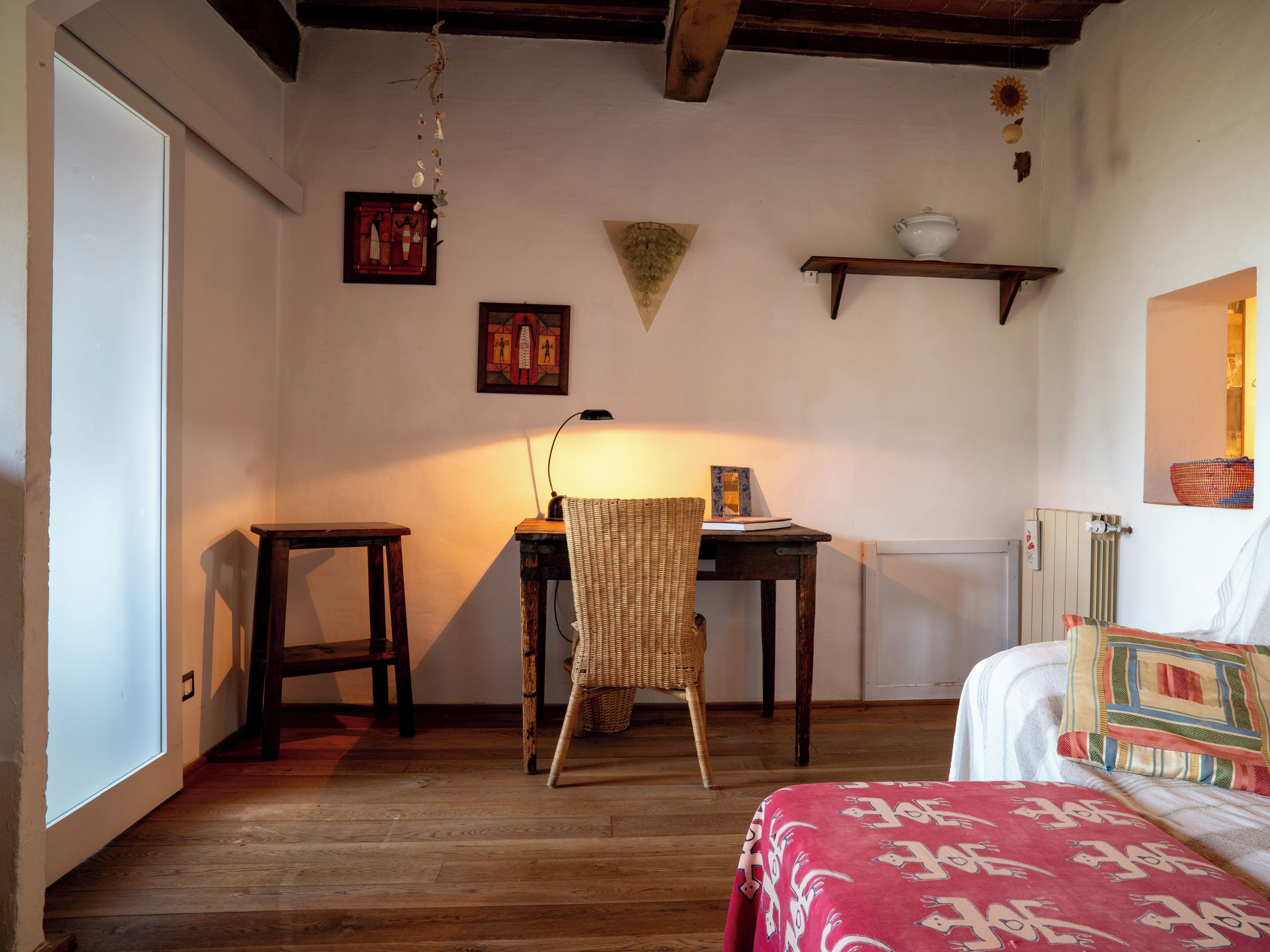A room with a wooden floor, a bed with patterned bedding, a desk with a lamp, a chair, and wall-mounted decorations.