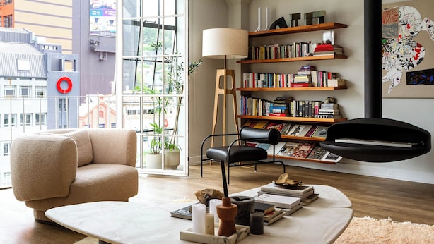 Uma sala de estar moderna com um sofá, uma mesa de centro com livros e velas, uma estante e uma vista da cidade através de grandes janelas.