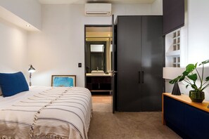 A bedroom with a bed, bedside lamp, a framed picture, a dark wardrobe, and a plant on a wooden side table. - Westwood (Cape Town)