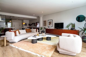 A modern living room with a sofa, coffee table, and a TV. There is a kitchen visible in the background.