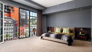 Quarto moderno com cama grande, vista da cidade através de janelas do chão ao teto e paisagem urbana visível.