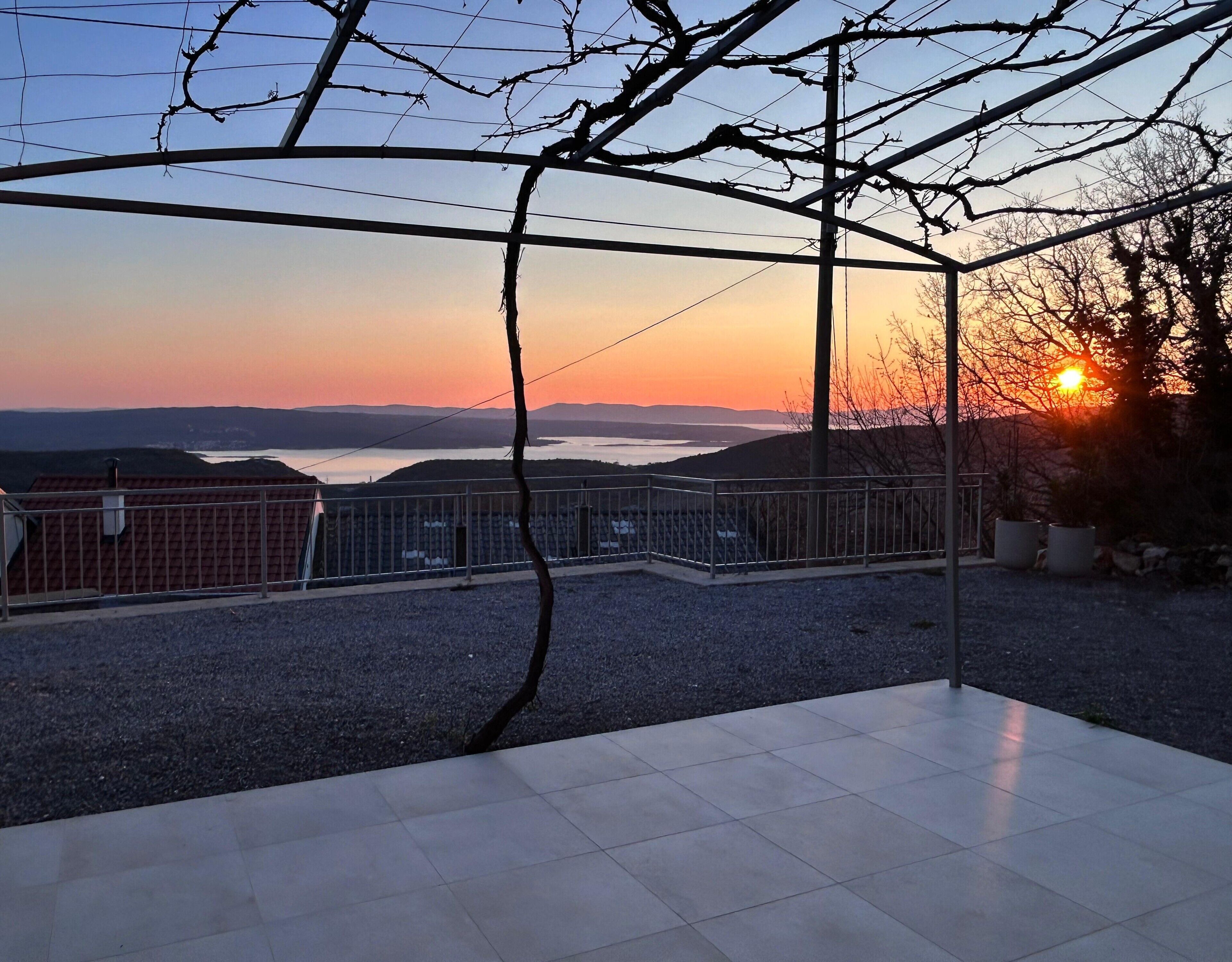 Une terrasse offrant une vue sur un coucher de soleil se reflétant sur une étendue d’eau, entourée d’une clôture en métal et dotée d’un sol carrelé.
