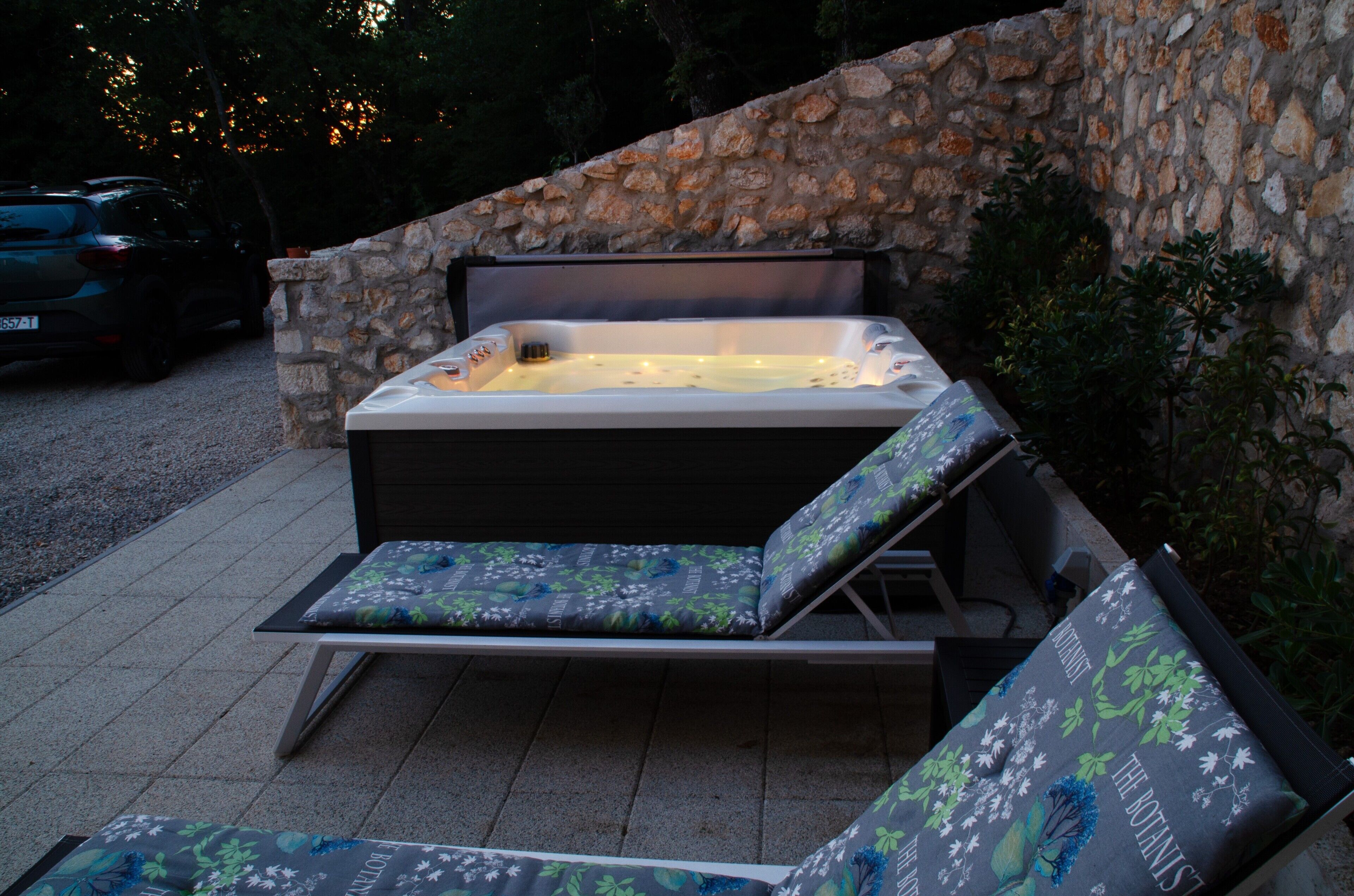 A hot tub with two lounge chairs, set against a stone wall, outdoors.