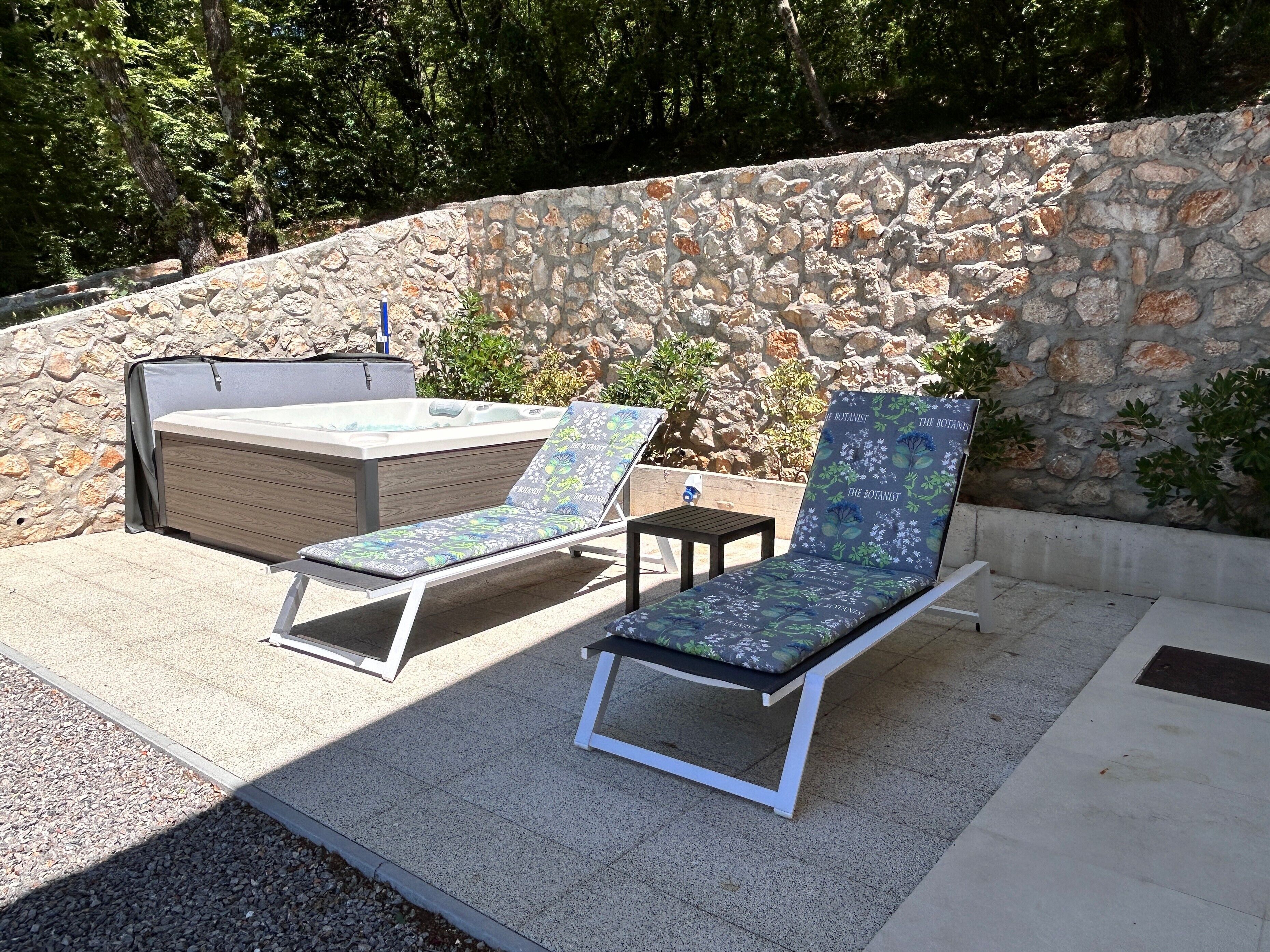 A stone wall, two lounge chairs, and a hot tub in an outdoor setting.