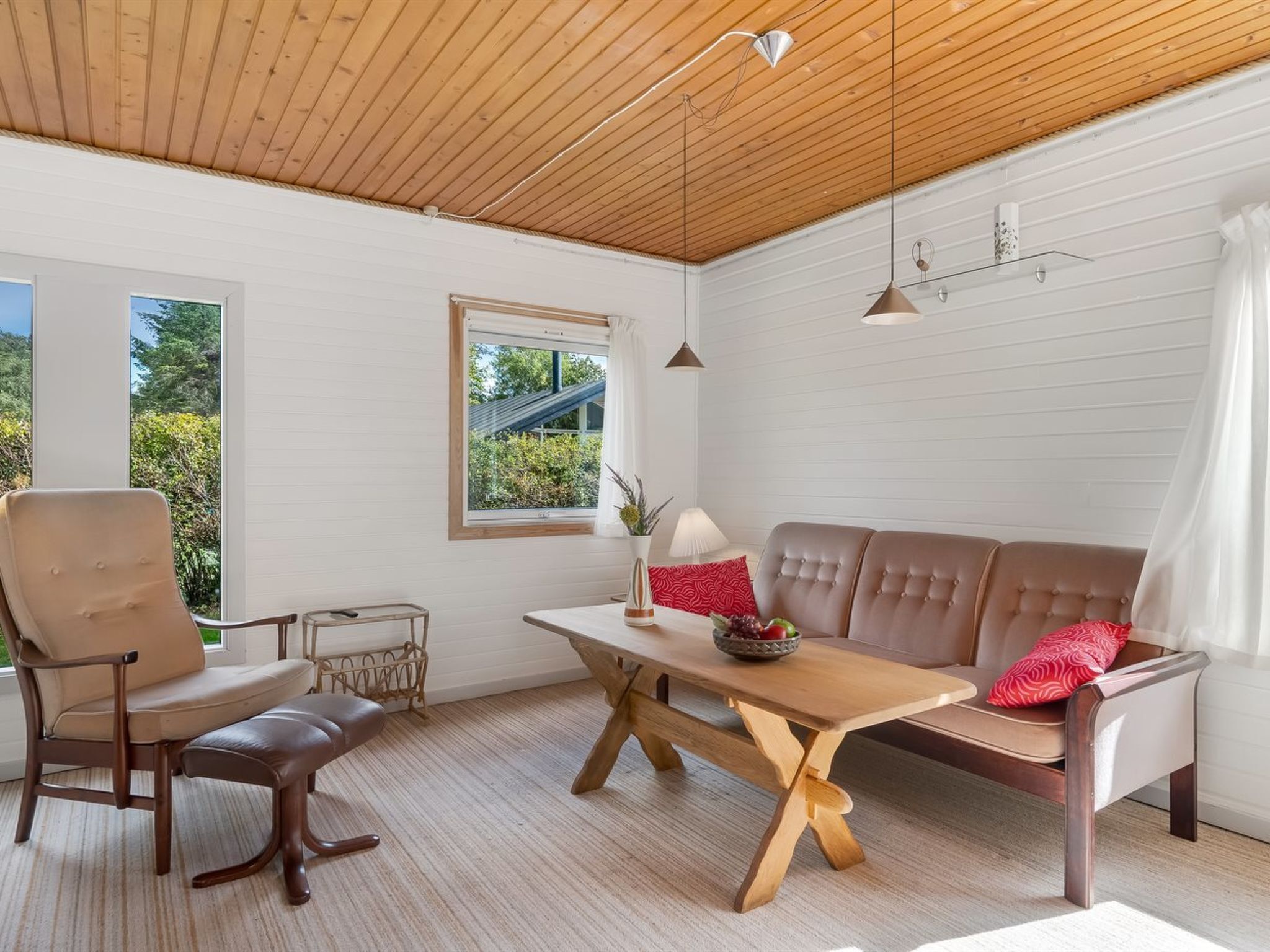 Un intérieur moderne avec un plafond en bois, des murs blancs, un canapé, un fauteuil, une table et une fenêtre donnant sur un paysage verdoyant.