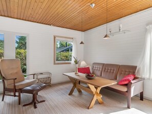 Un interior moderno con techo de madera, paredes blancas, un sofá, una silla, una mesa y un ventanal con vistas a zonas verdes.
