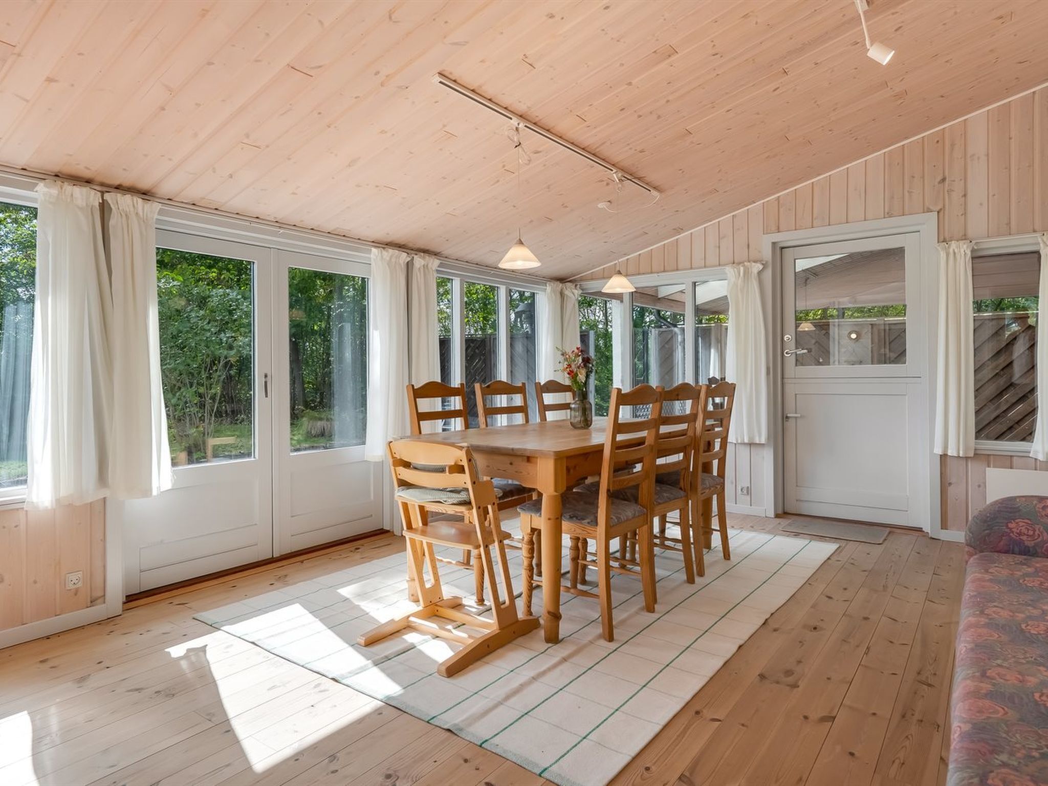 A wooden dining table with six chairs, a vase with flowers, and a view of the outdoors through large windows.