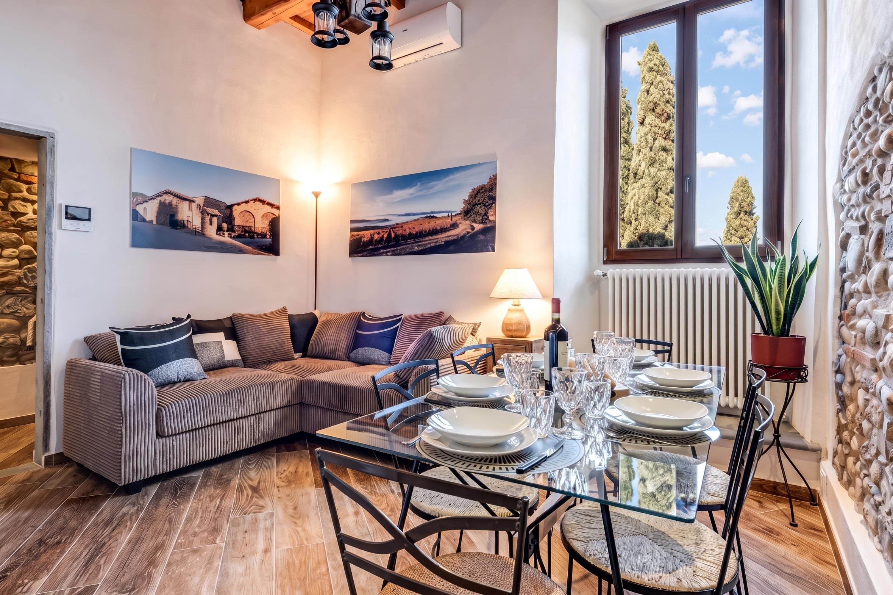 A cozy living room with a sofa, a dining table set for four, and two large windows with a view of trees.