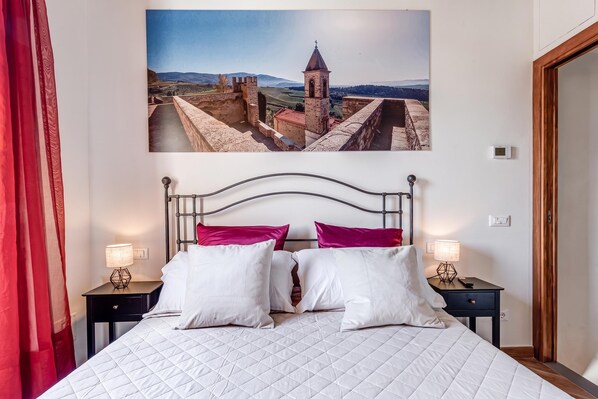 A bed with a white quilted bedspread, two white pillows, and two red pillows, flanked by two bedside tables with lamps, in a room with a large landscape painting on the wall. - Luxury Chianti Road - 3BR Free Parking (San Casciano in Val di Pesa)
