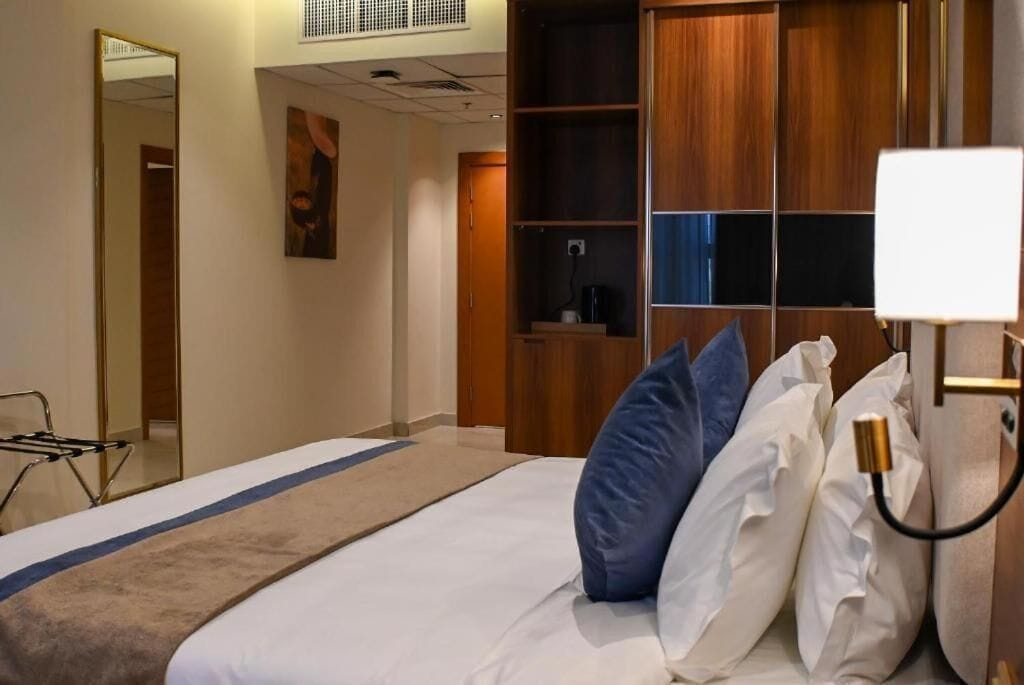 A hotel room with a bed, a bedside table with a lamp, a wardrobe, a mirror, and a wall-mounted shelf.